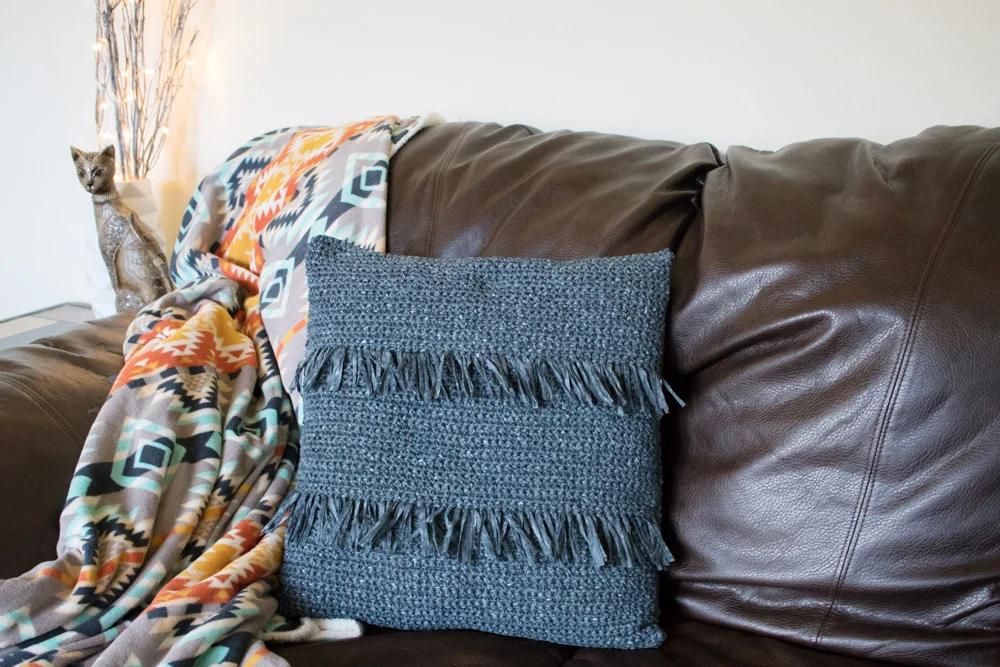 Gray crochet throw pillow with two horizontal rows of fringe, propped on a brown leather sofa with a blanket