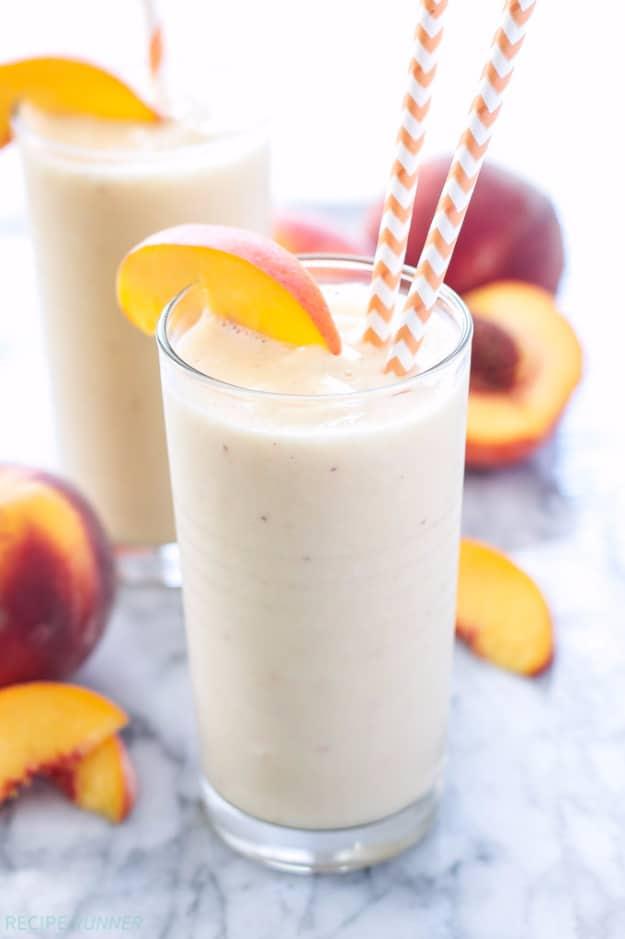 Two tall glasses of creamy peach green tea smoothie with peach slices on the rims and orange striped straws on marble