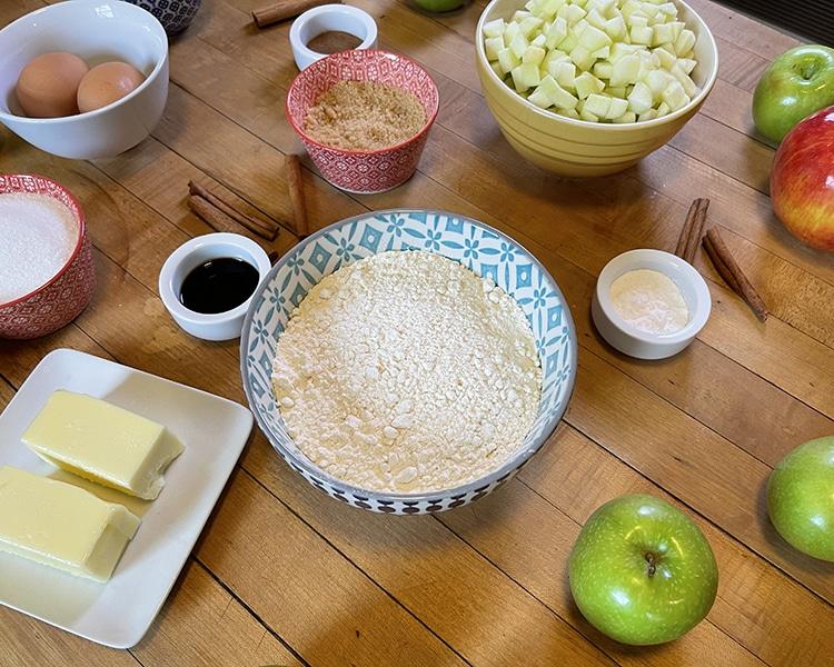 Apple Recipes - blue patterned bowl of flour surrounded by two butter sticks on a white plate, diced apples, brown sugar, vanilla, cinnamon sticks, whole apples and eggs