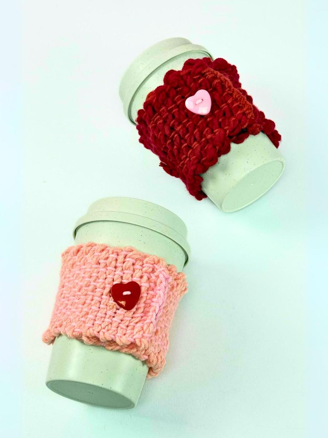 Two woven yarn cup cozies, one red and one pink, each with a heart button, wrapped around pale green travel cups