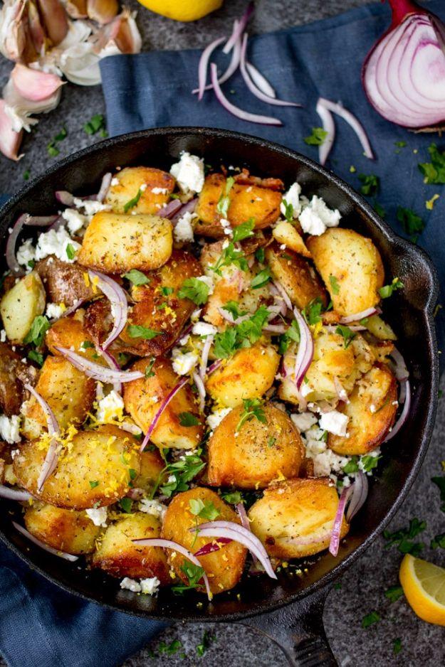 Cast iron skillet of crispy Greek potatoes with red onion slices, crumbled feta, parsley and lemon wedges