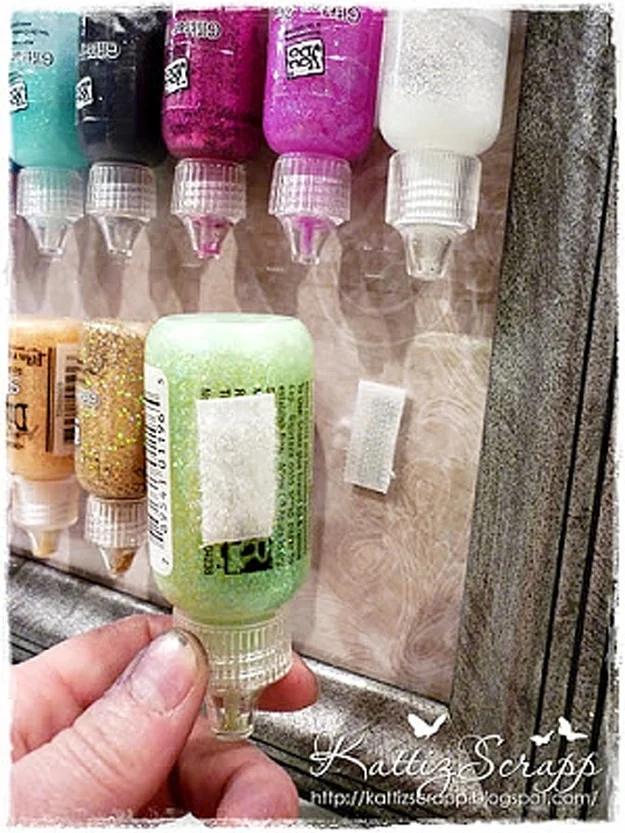 Glitter glue bottles mounted upside down on the glass of a framed board with velcro dots, a hand lifting one off
