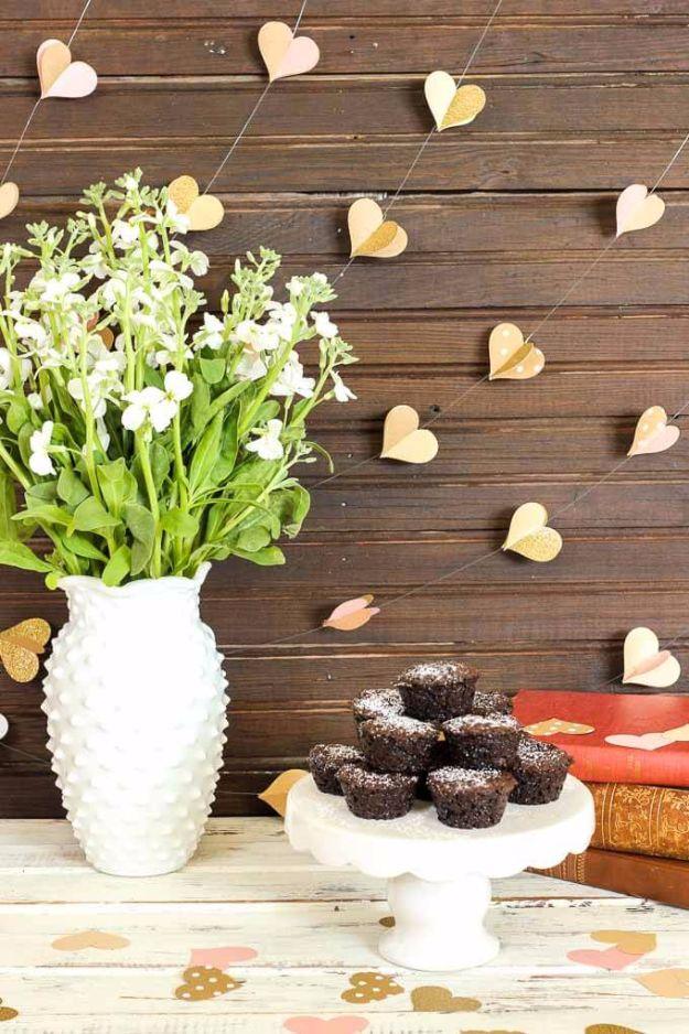 Dark wood wall hung with strings of small gold paper hearts above a white vase of flowers and a plate of brownies