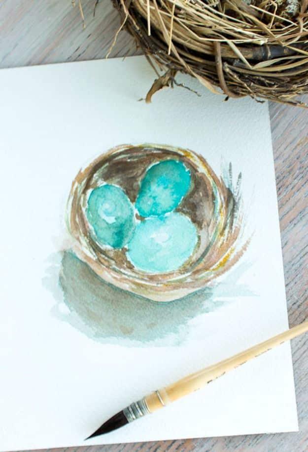 Watercolor nest printable with three turquoise eggs beside a paintbrush and a real nest on weathered wood