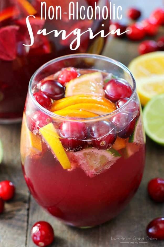 Glass of red non alcoholic sangria with orange slices and cranberries beside cut lime and lemon on wood