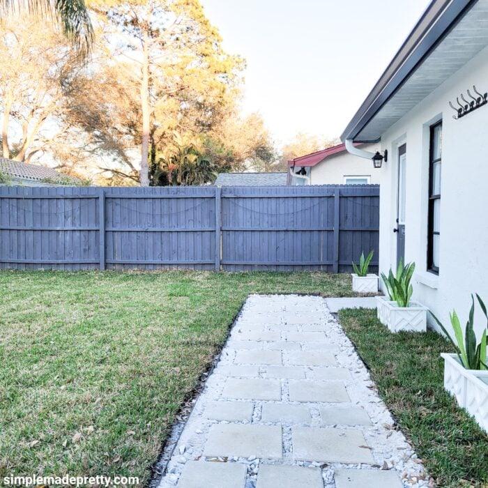 Wide pale paver walkway running alongside a white house and a blue fence, cutting across a green lawn