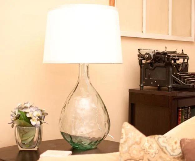 Glass jug table lamp with a white shade on a desk beside a vintage typewriter and a small vase of flowers