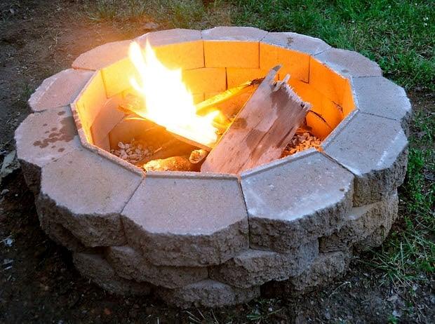 Round fire pit built from angled concrete blocks with a log fire burning inside, set on grass in a backyard
