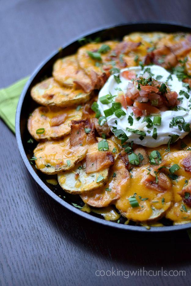 Irish potato nachos in a black skillet, sliced potatoes under melted cheddar with bacon, sour cream and chives