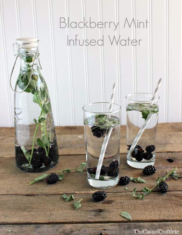 Blackberry mint infused water in a swing top bottle with two glasses of it, striped straws and loose berries on wood