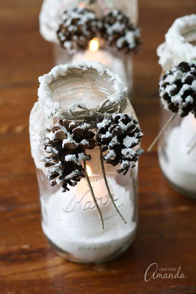 Glass jar luminary with lace around the rim, a twine bow and snow textured pine cones glowing from a candle inside
