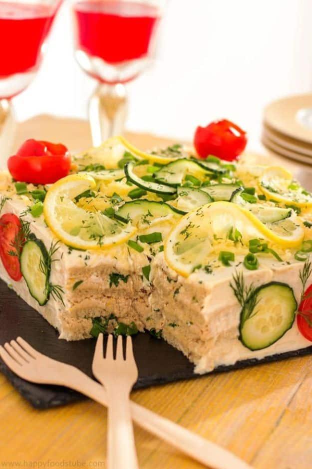 Savory tuna sandwich cake frosted white and topped with lemon slices, cucumber, dill and cherry tomatoes