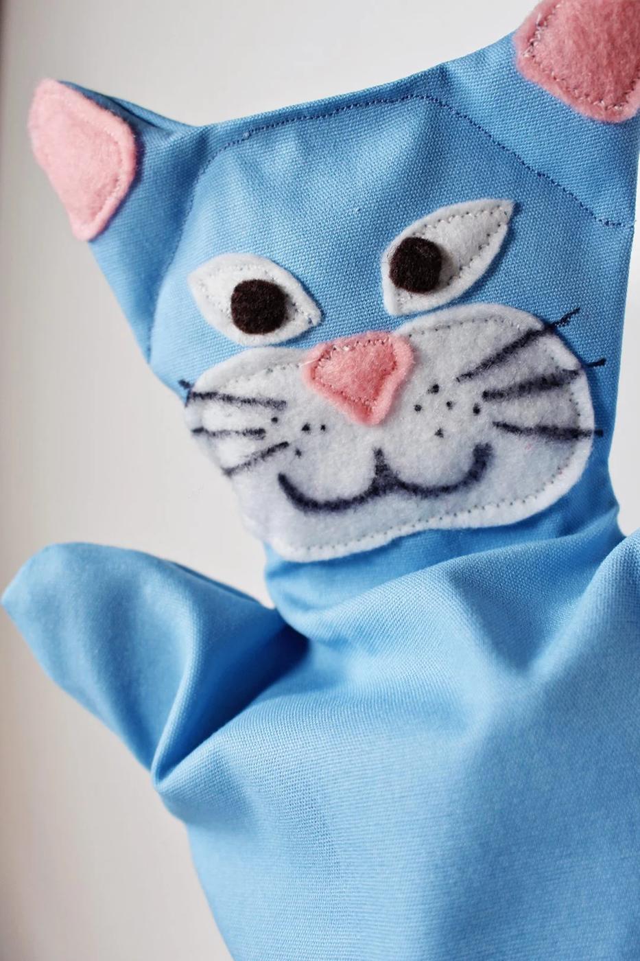 Finished blue fabric cat hand puppet with pink felt ears, white muzzle and stitched eyes standing against a white wall