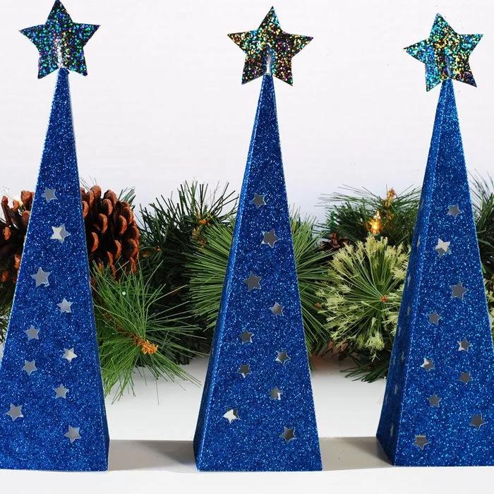 Three tall blue glitter paper pyramid tree lanterns with star cutouts and glitter star toppers in front of greenery