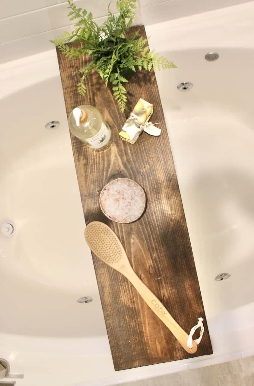 Stained pine board bathtub tray across a white tub holding a fern, body wash, a bar of soap, bath salts and a bamboo brush