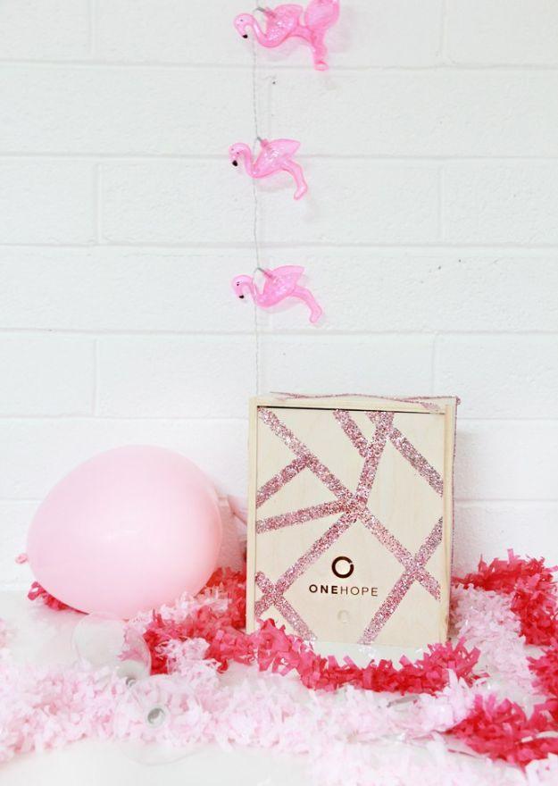 Small wooden gift crate with a criss cross pattern of pink glitter lines, staged with a pink balloon and paper shred