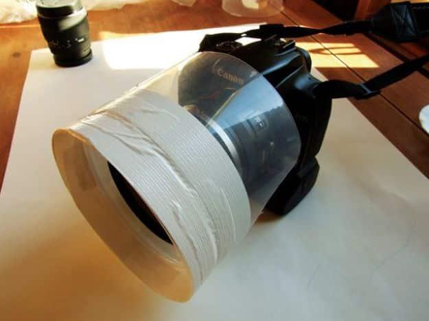 Camera rain guard, a translucent plastic ring taped around the front of a Canon DSLR lens on white paper on a wooden table