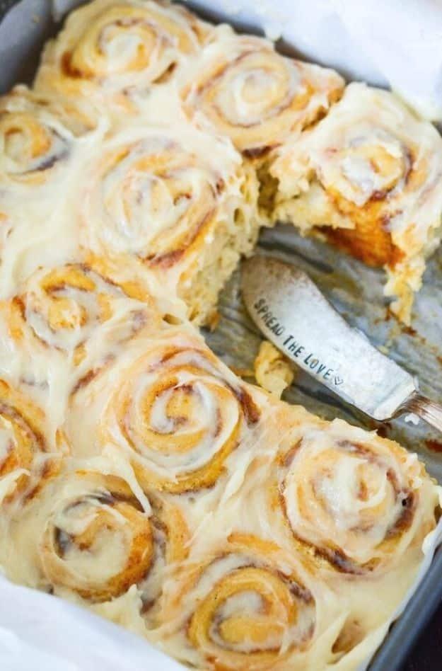 Pan of pumpkin cinnamon rolls under thick cream cheese icing, one roll gone and a spreader left in the gap