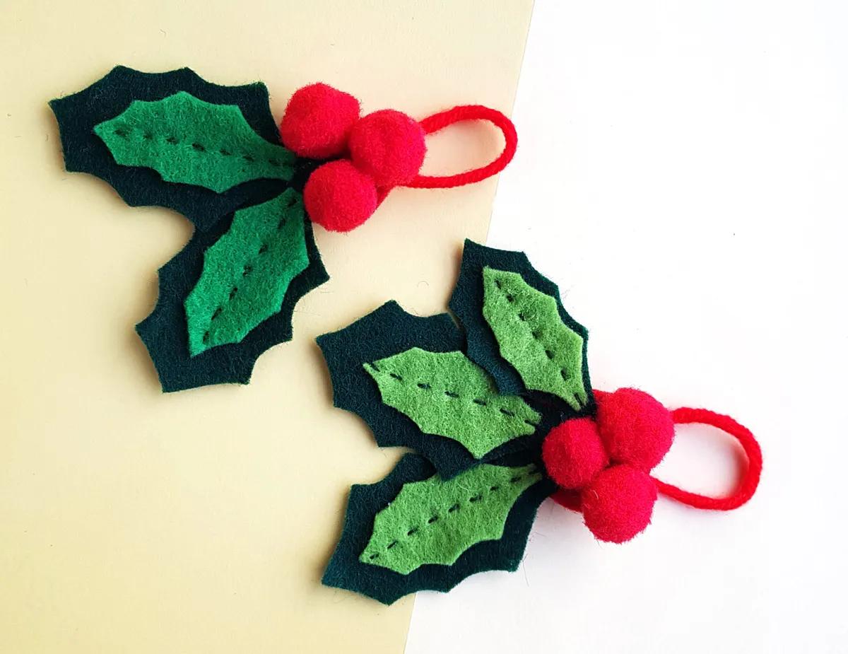 Two felt holly ornaments with layered green leaves, stitched veins, red pom pom berries and red yarn loops on cream paper