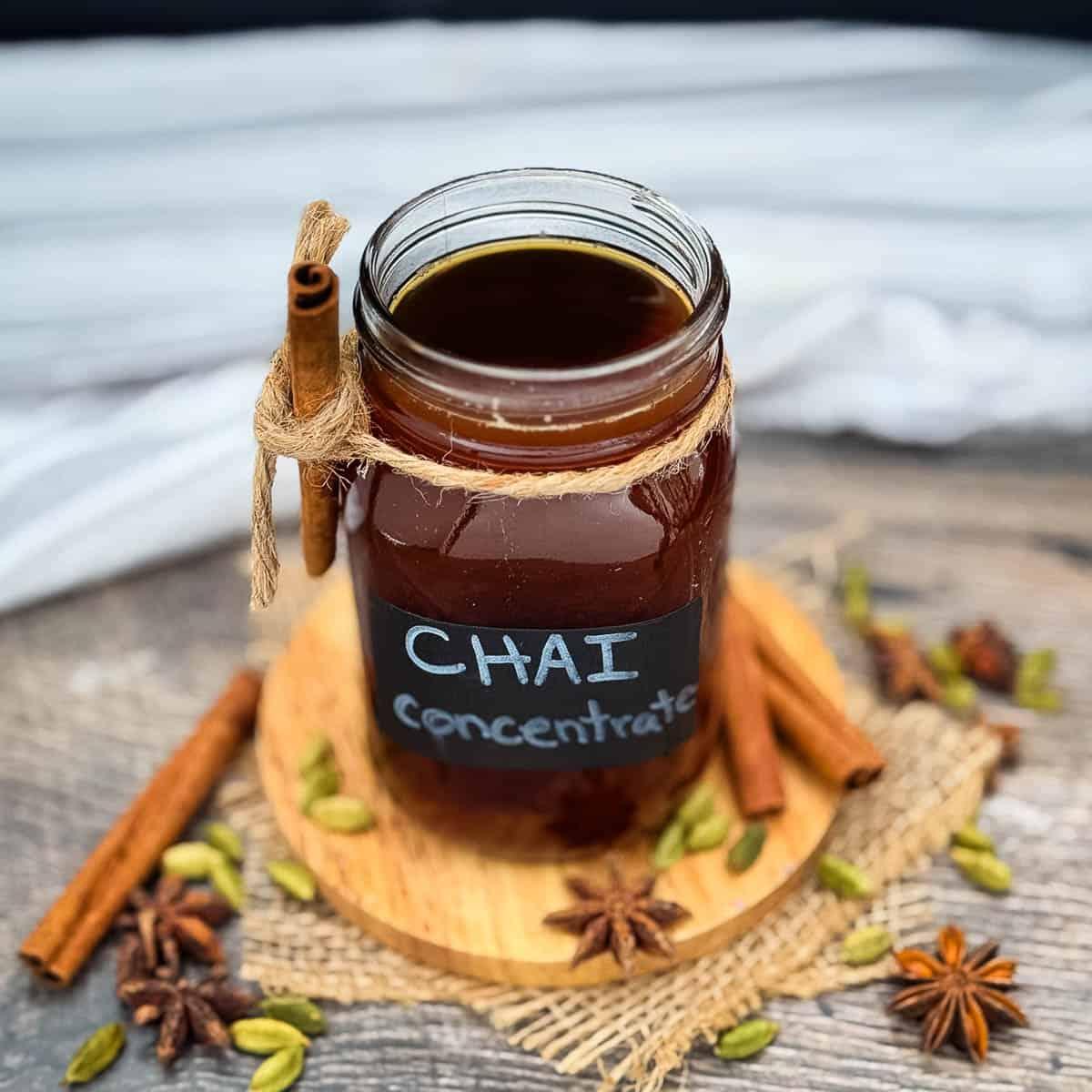 Glass jar of dark amber chai concentrate with a cinnamon stick tied at the neck, on a wood slice with star anise and cardamom