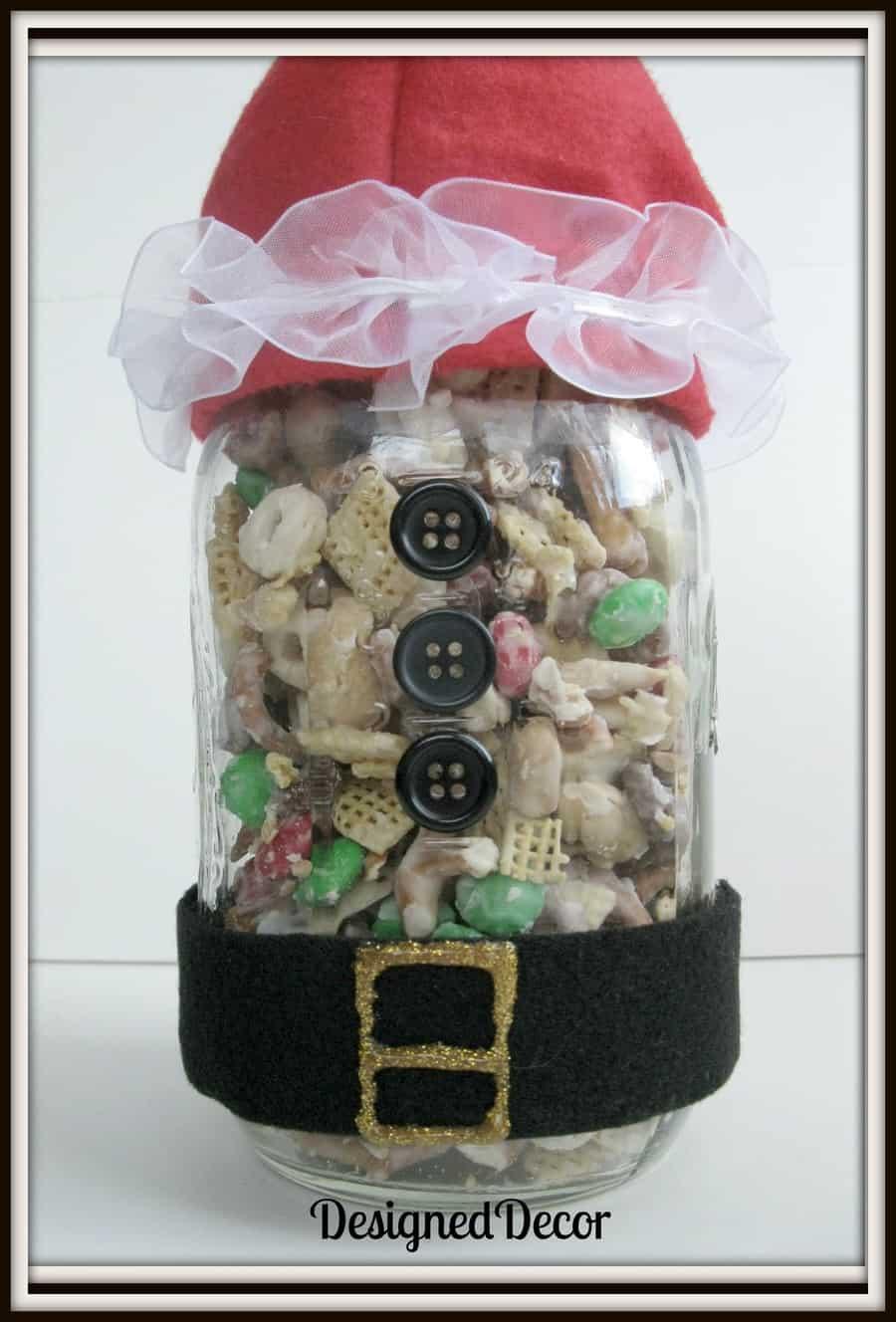 A glass jar of Christmas snack mix dressed as Santa with a black felt belt, buttons and a red felt hat