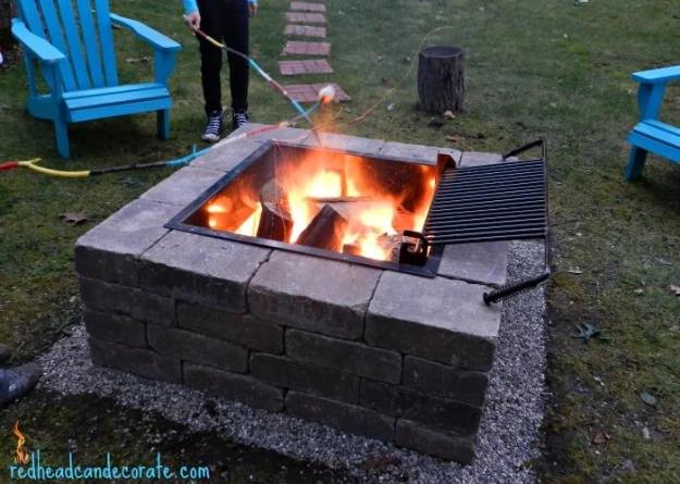 Square DIY firepit kit of stacked gray block with a swing-out steel grill grate over burning logs and blue chairs