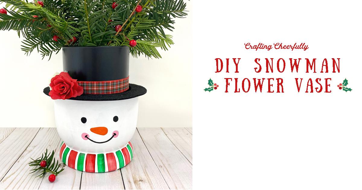 A DIY snowman vase painted white with a black felt hat, ribbon scarf and painted face, holding red flowers and greenery