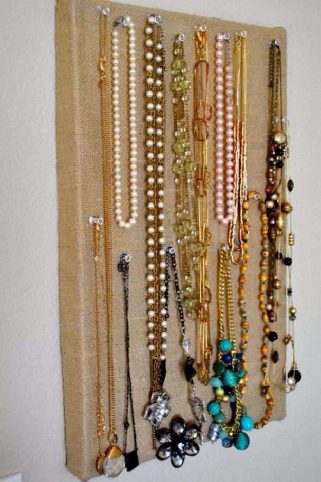 Boot box lid covered in burlap, hung on a wall and holding rows of necklaces on small tacks and nails