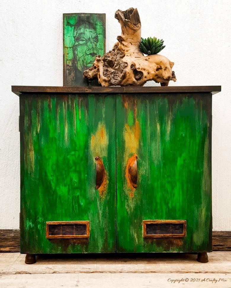 Melamine cupboard painted in vivid green with rust colored drips and a dark wood top, styled with driftwood and a succulent