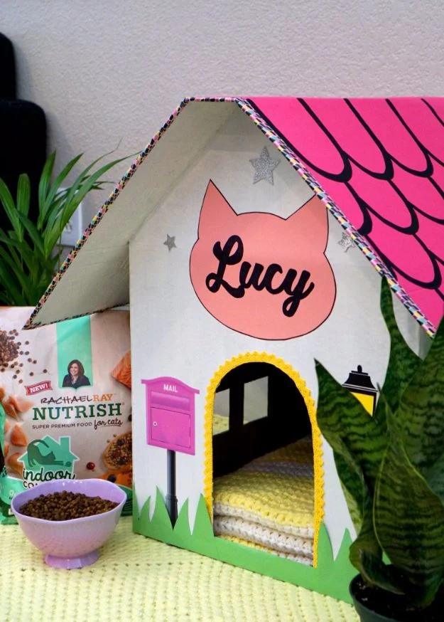 Cardboard craft cat house painted as a white cottage with a pink scalloped roof, arched door and a name sign