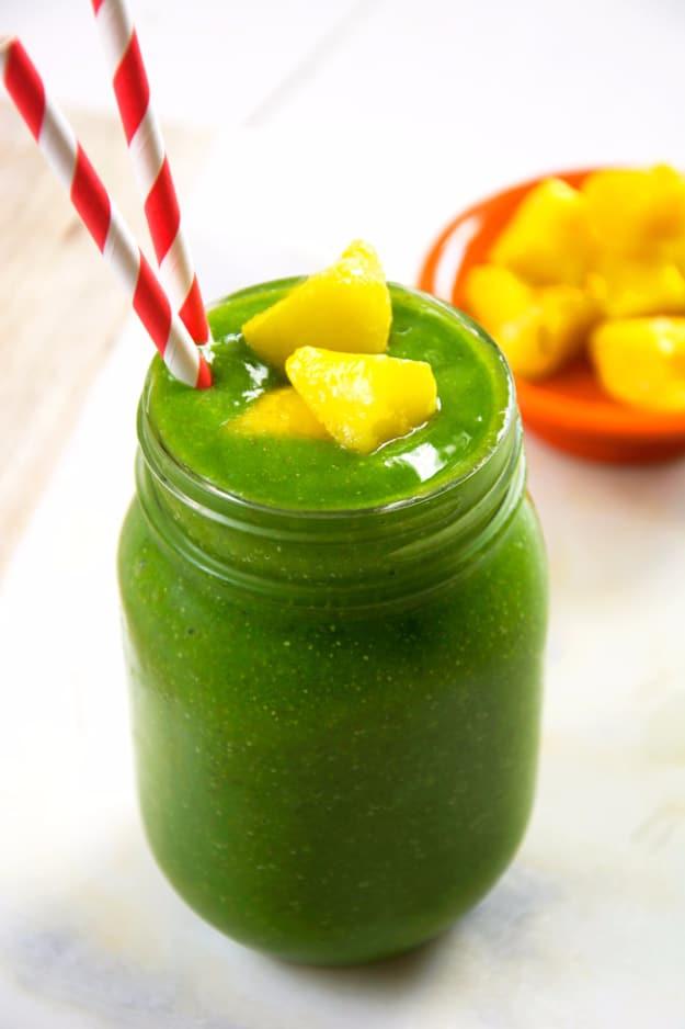 Bright green cucumber tropical smoothie in a mason jar topped with golden mango chunks and two red straws