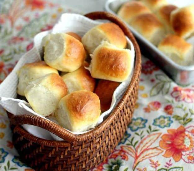 Basket of golden homemade dinner rolls lined with a cloth beside a baking dish of more rolls on a floral cloth