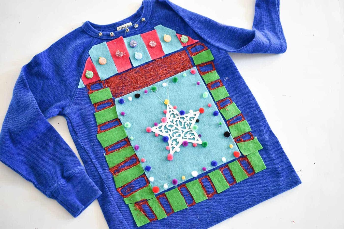 A blue kids sweatshirt covered in felt squares, green felt tabs, felt ornaments, pom poms, a lace snowflake and glitter paint