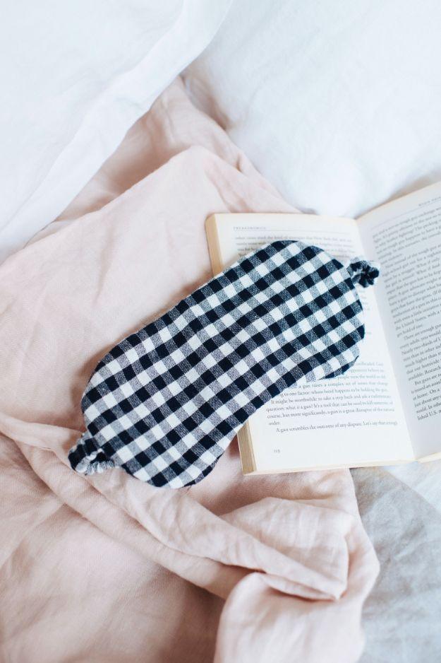 Navy gingham linen eye mask with an elastic strap resting on a pale pink linen blanket beside a book
