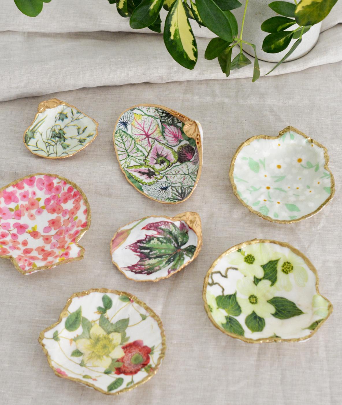 Six scallop shells decoupaged with floral and botanical napkin prints and gold painted rims on white linen with greenery