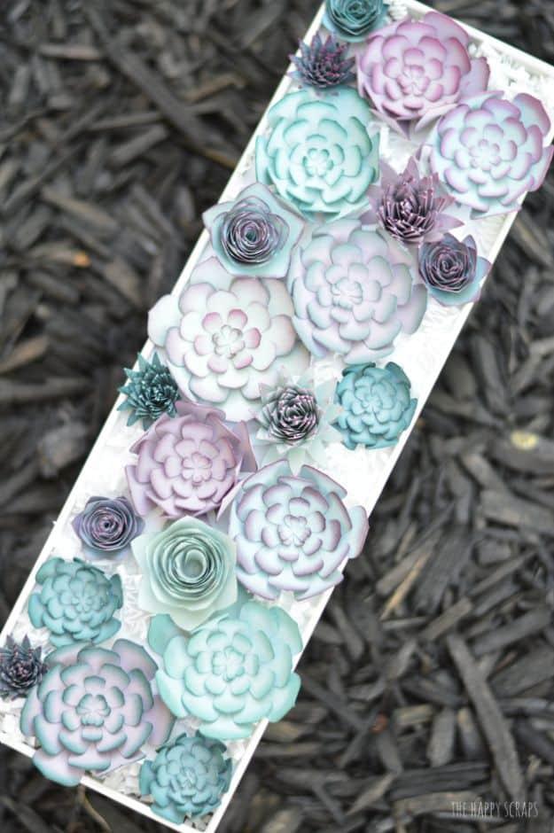 Long white tray filled with layered paper succulents in teal, lilac and sage, photographed from above on dark mulch