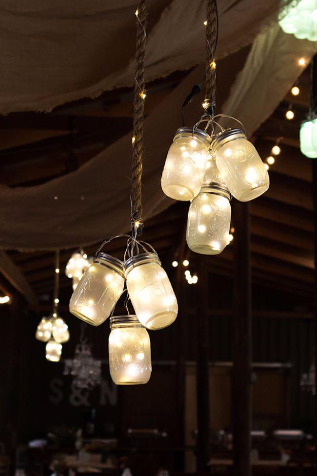 Cluster of glowing frosted mason jars hung on twisted cords from a dark barn ceiling above a lit reception