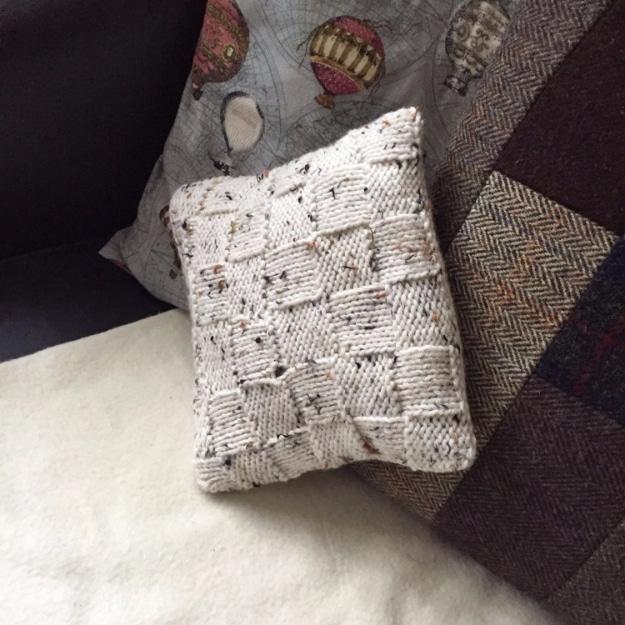 Cream flecked basket weave knitted cushion cover on a patterned pillow against a dark sofa and tartan blanket