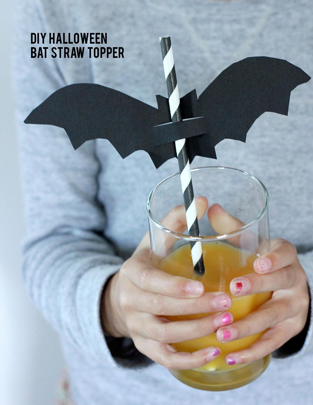 Black cardstock bat straw topper threaded onto a striped straw above a glass, with a DIY Halloween headline and hands