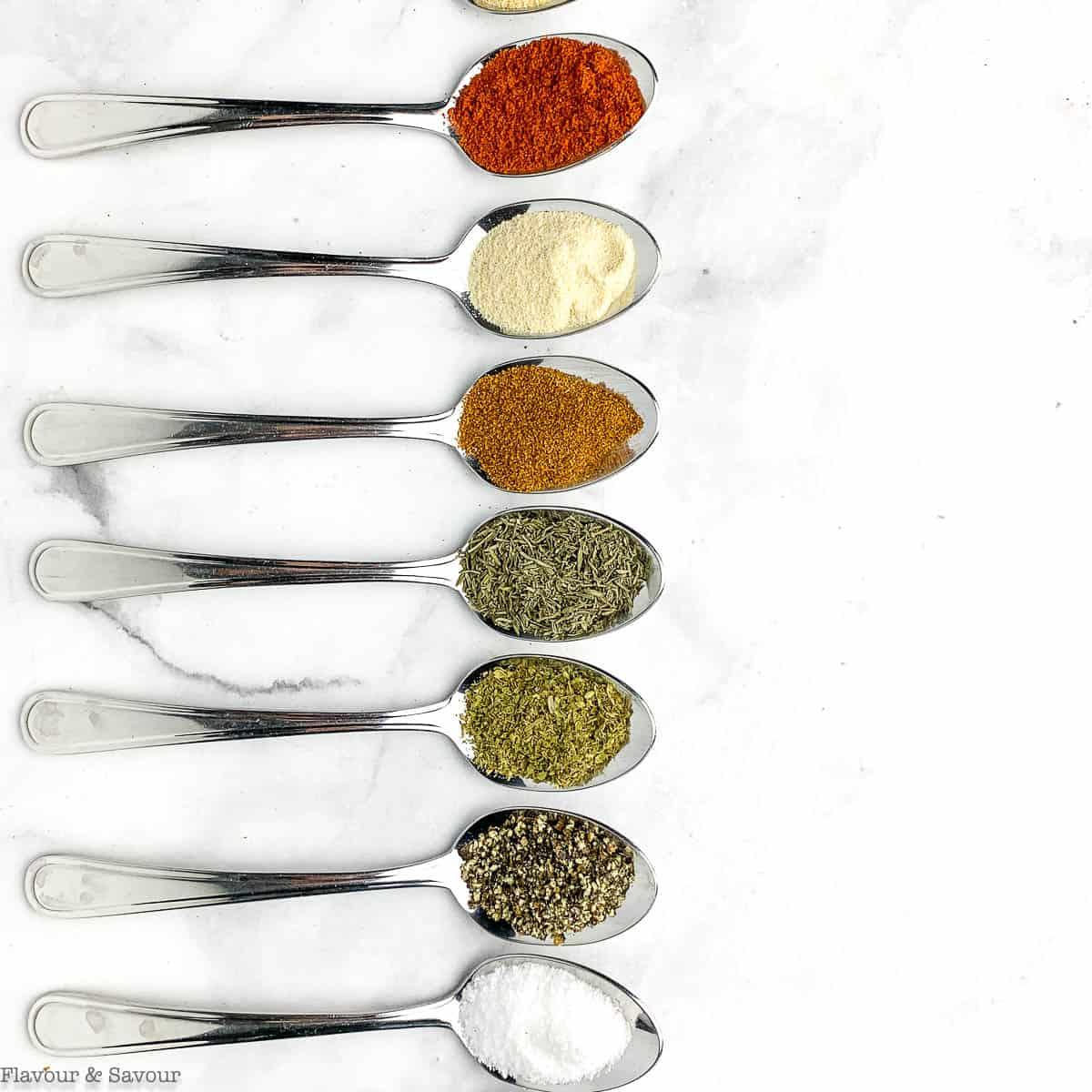 Row of seven silver spoons on white marble, each holding a different spice including paprika, garlic powder and dried herbs