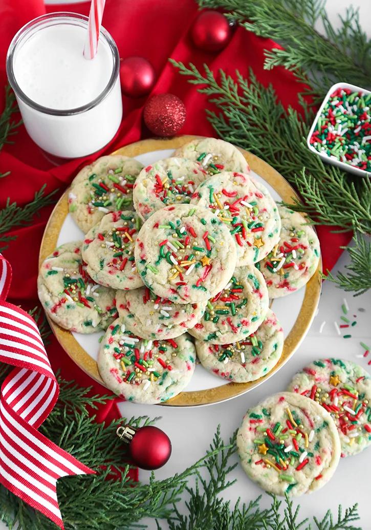 A plate of soft sugar cookies loaded with red, green and white sprinkles and gold stars beside a glass of milk and evergreens