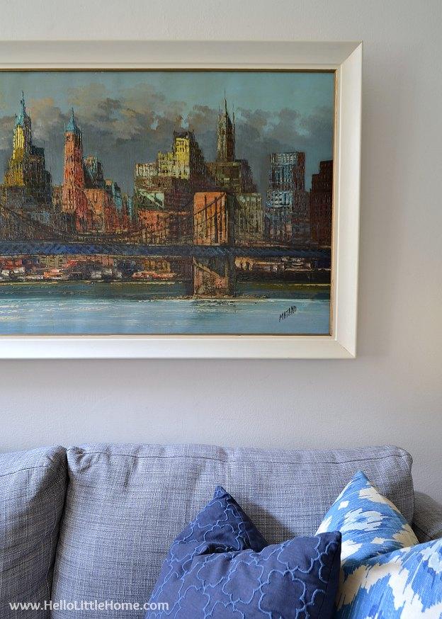 Large painting in a freshly painted white frame hanging on a wall above a gray sofa with blue and white pillows