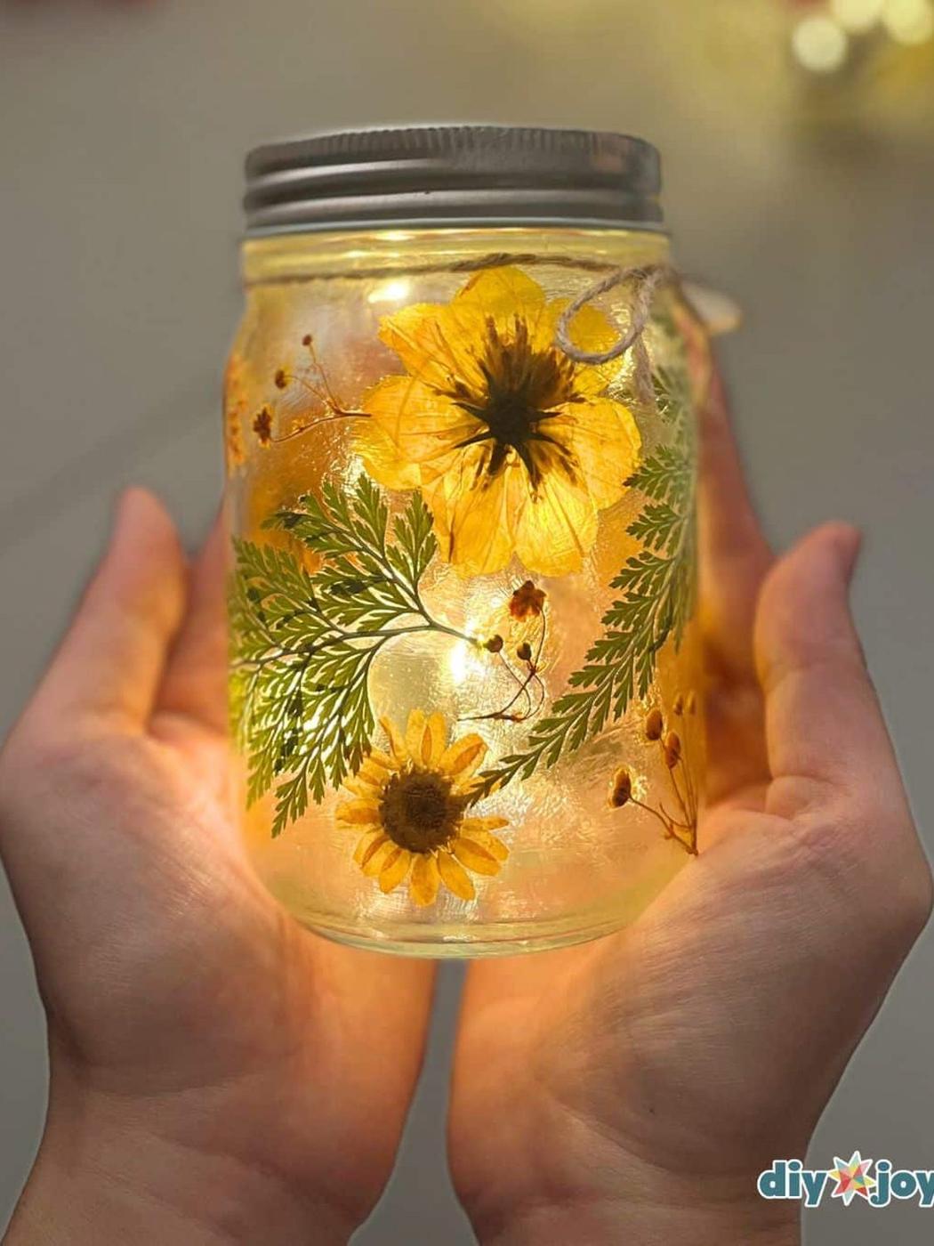 A glass mason jar covered in pressed flowers and leaves with fairy lights inside making the petals glow in a dark room