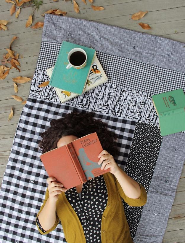 Quilt of black and white gingham, stripe and dot panels on decking with a woman lying on it holding a book over her face