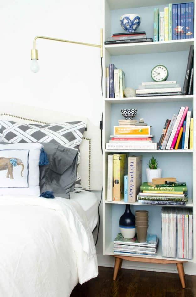White bookcase with pale blue painted back and angled wooden legs beside a bed, brass swing arm sconce and books