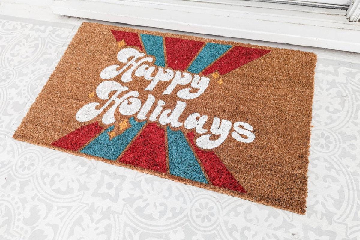Coir doormat stenciled with Happy Holidays in white script over red and turquoise stripes on a front porch