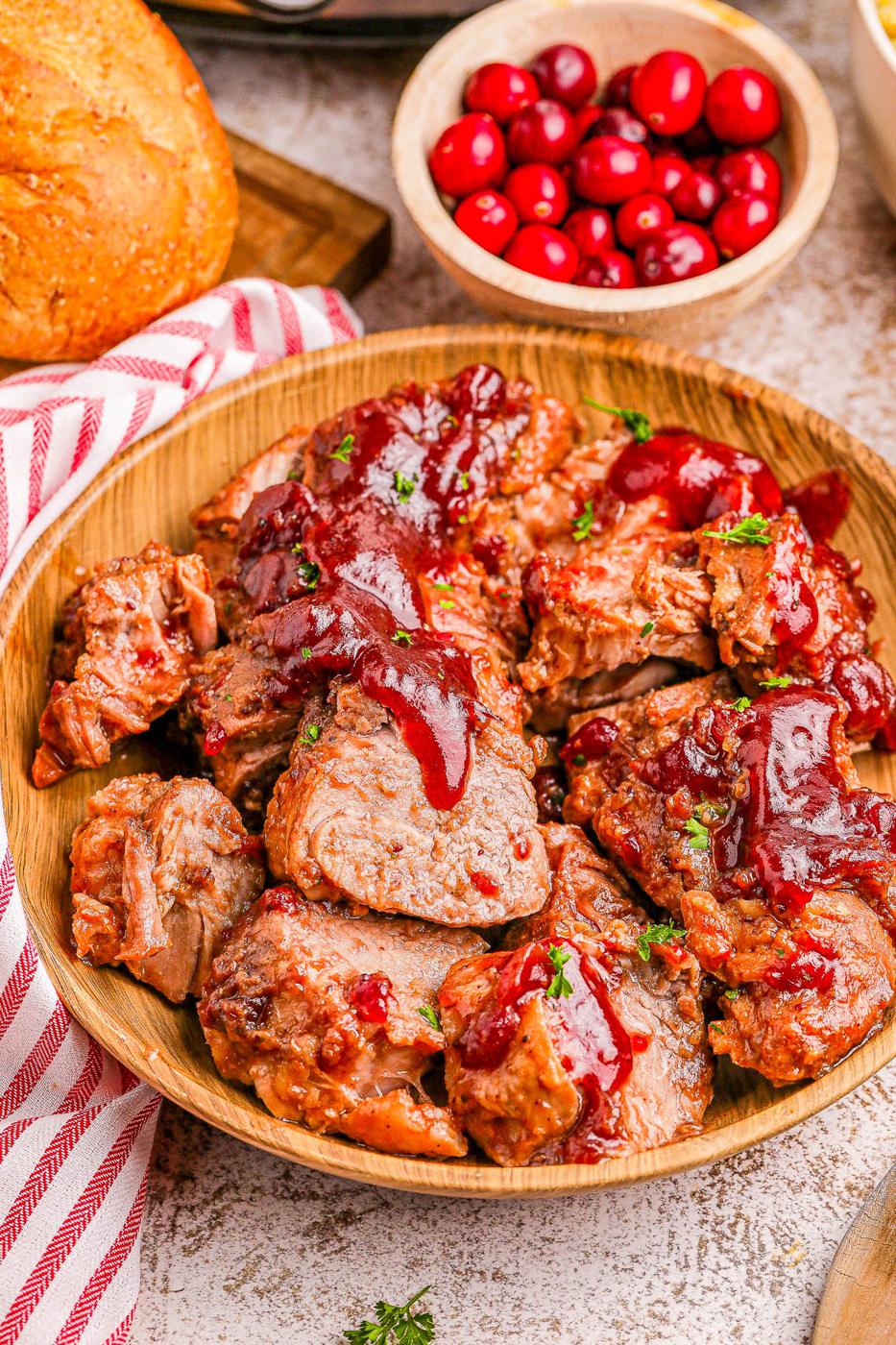 Sliced pork tenderloin covered with cranberry glaze in a wooden bowl with cranberries and bread behind