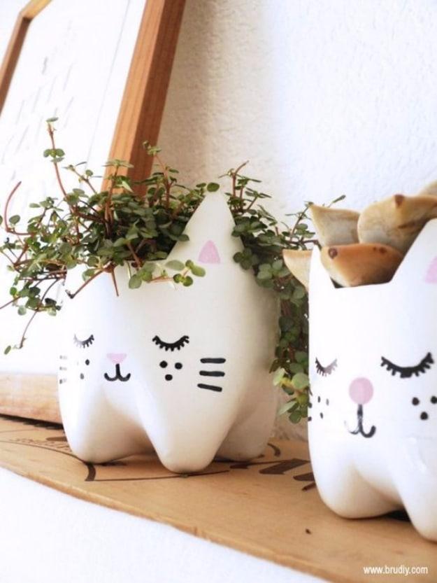 Two white plastic bottle planters painted as sleeping cat faces with pink ears and noses, holding small trailing green plants