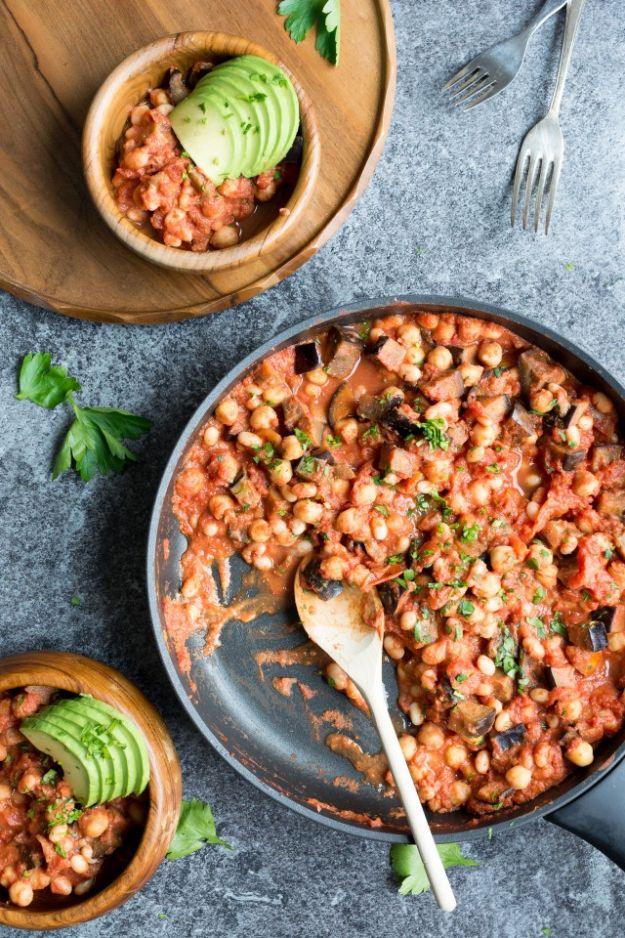 Creamy vegan bean and eggplant casserole in a skillet with a wooden spoon and side bowls topped with avocado