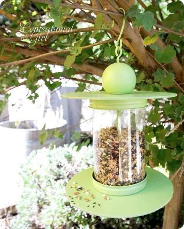 Green painted recycled bottle bird feeder with round wood disks and a ball finial, hanging in a leafy tree